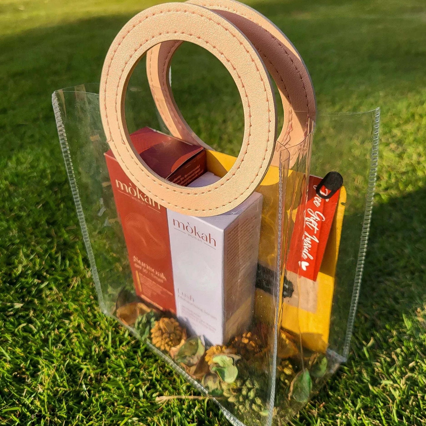 Mòkah Skin Love Duo in a clear chic pouch containing skincare products and hot chocolate, set on grass.